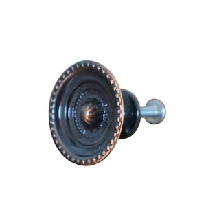 May include: A decorative cabinet knob with a dark bronze finish. The knob features a circular design with a beaded edge and a central, textured pattern. The stem and handle are black and silver, respectively.