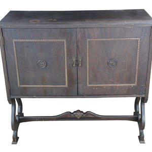 May include: A dark brown wooden cabinet with two doors, brass hardware, and decorative trim. The cabinet has a curved top and is supported by a decorative base. The doors feature a square design with a circular emblem.