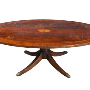 May include: A wooden oval coffee table with a decorative inlay in the center of the top. The table has a pedestal base with four legs.