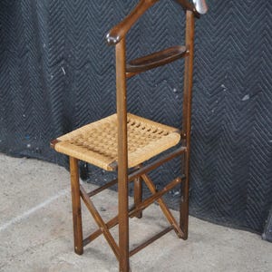 May include: A vintage wooden valet chair with a woven seat and a built-in coat hanger. The chair has a dark brown finish and a small shelf. The coat hanger has a number 1 on it. The chair is designed to hold clothing.