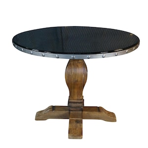 May include: A round table featuring a dark, textured top with a silver-toned metal rim accented by evenly spaced rivets. The table is supported by a central, carved wooden pedestal base that transitions into a four-legged cross-shaped foundation.