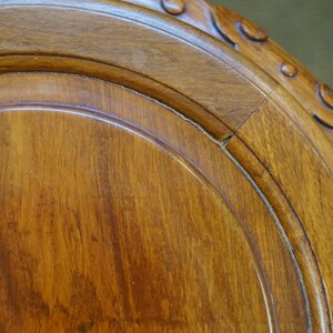 May include: Close-up of a section of a wooden table. The wood has a warm, reddish-brown hue with visible grain patterns. The edge features carved decorative elements. The table appears to be antique or vintage.