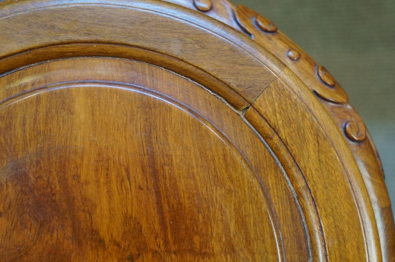May include: Close-up of a section of a wooden table. The wood has a warm, reddish-brown hue with visible grain patterns. The edge features carved decorative elements. The table appears to be antique or vintage.