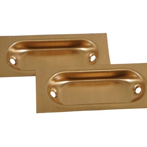 May include: Two brass-colored recessed pulls. Each pull has a rectangular backplate with a rounded, oval-shaped handle and two circular holes. The pulls are likely for a cabinet or drawer.