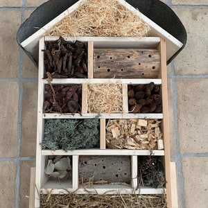 May include: A multi-compartment wooden insect hotel with a peaked roof. The structure contains various natural nesting materials including wood shavings, pine cones, dried straw, moss, bark, and two logs with drilled holes for solitary bees.