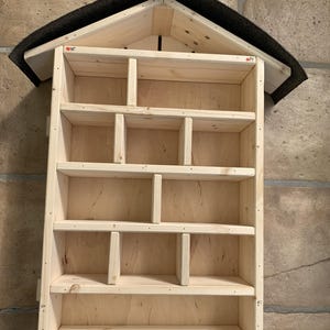 May include: A light-colored wooden insect hotel featuring a gabled roof covered in dark grey roofing felt. The structure is divided into multiple rectangular compartments of varying sizes by internal wooden dividers, creating a grid-like storage space.