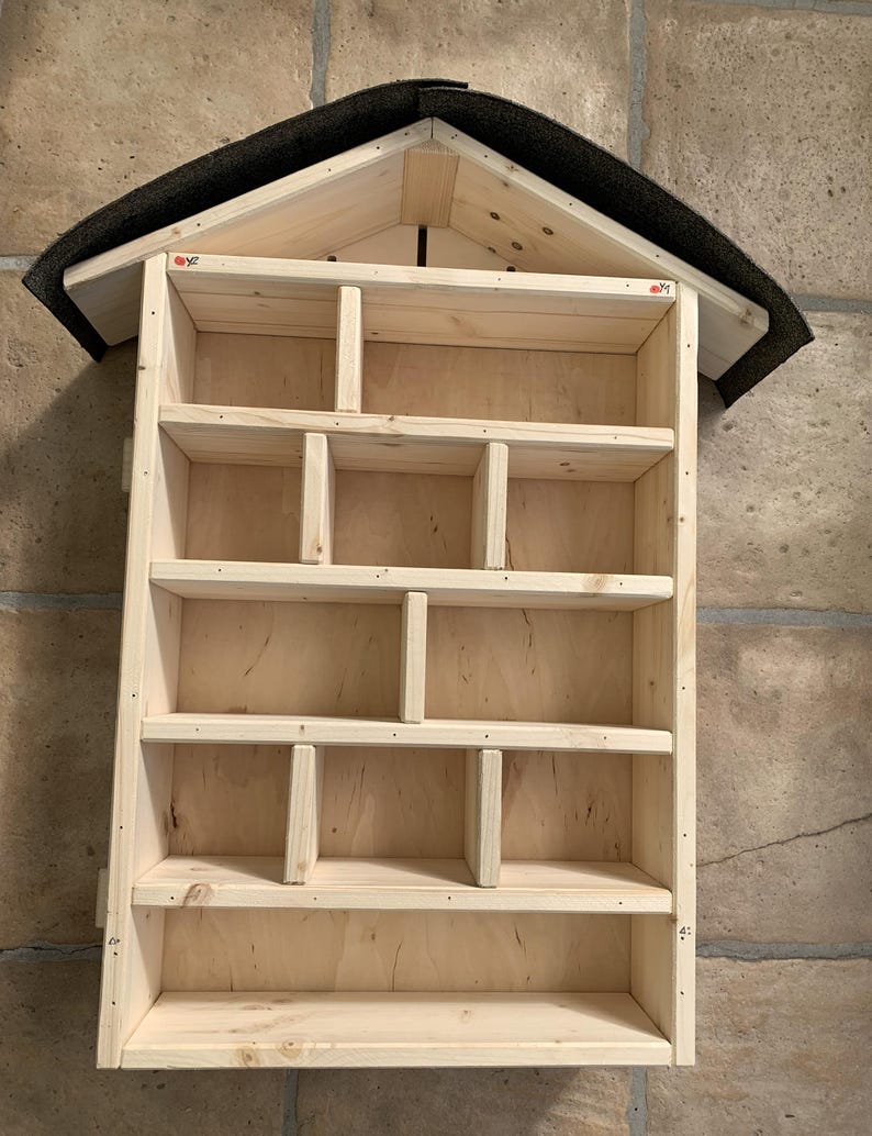 May include: A light-colored wooden insect hotel featuring a gabled roof covered in dark grey roofing felt. The structure is divided into multiple rectangular compartments of varying sizes by internal wooden dividers, creating a grid-like storage space.