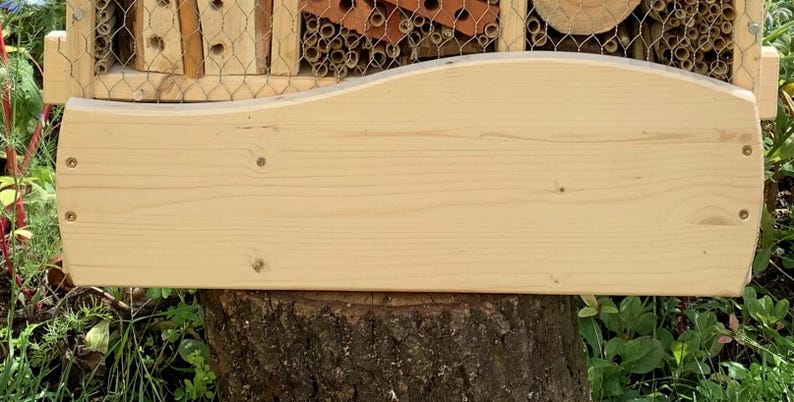 May include: A rectangular light-colored wooden board with a smooth, curved top edge and four small metal screws visible on the front surface. The board features a natural wood grain pattern and is positioned horizontally in front of a wooden insect hotel.