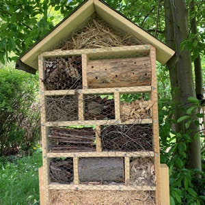 May include: A wooden insect hotel with a peaked roof, featuring multiple compartments filled with natural materials like pinecones, drilled logs, twigs, wood chips, and straw, all secured behind a fine metal wire mesh grid for protection.