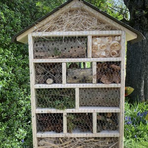 May include: A rectangular wooden insect hotel with a peaked roof, featuring multiple compartments filled with natural materials like wood logs with drilled holes, pine cones, wood shavings, twigs, and dried moss, all secured behind a wire mesh screen.