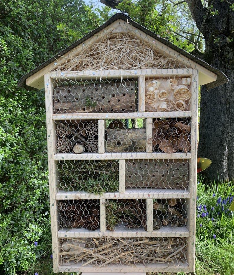 May include: A rectangular wooden insect hotel with a peaked roof, featuring multiple compartments filled with natural materials like wood logs with drilled holes, pine cones, wood shavings, twigs, and dried moss, all secured behind a wire mesh screen.