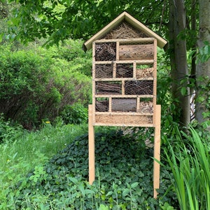May include: A wooden insect hotel with a peaked roof, supported by two vertical legs. The structure is divided into multiple rectangular compartments filled with natural materials like pinecones, twigs, wood logs, and straw, secured by wire mesh.