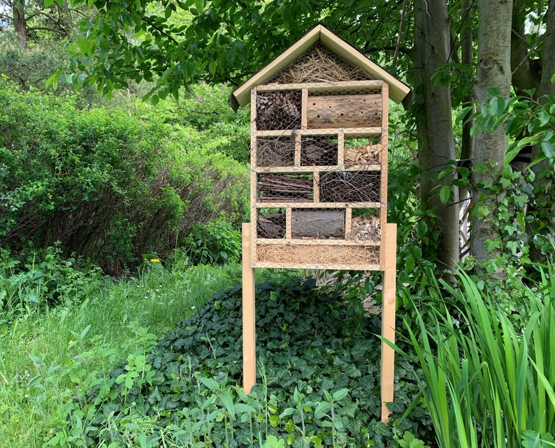 May include: A wooden insect hotel with a peaked roof, supported by two vertical legs. The structure is divided into multiple rectangular compartments filled with natural materials like pinecones, twigs, wood logs, and straw, secured by wire mesh.