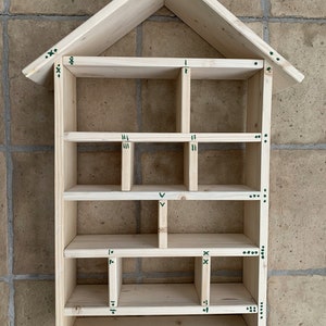 May include: A light-colored wooden wall shelf shaped like a house with a peaked roof. The interior is divided into multiple rectangular compartments of varying sizes by horizontal and vertical dividers. Small green hand-drawn markings are visible.
