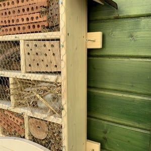 May include: A wooden insect hotel featuring multiple compartments filled with natural materials like terracotta bricks, hollow reeds, wood bark, and straw, all secured behind a protective wire mesh grid and mounted on a light-colored wooden frame.
