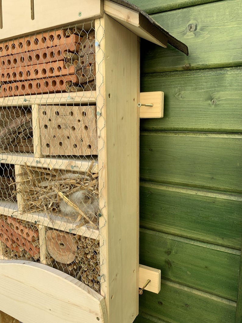 May include: A wooden insect hotel featuring multiple compartments filled with natural materials like terracotta bricks, hollow reeds, wood bark, and straw, all secured behind a protective wire mesh grid and mounted on a light-colored wooden frame.