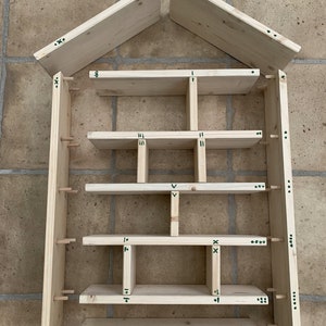 May include: An unassembled wooden house-shaped shelving unit consisting of light-colored pine boards, horizontal shelves, vertical dividers, and two roof panels. The components feature small green markings and wooden dowels for assembly.