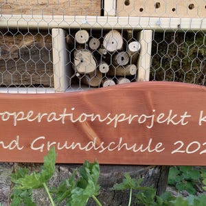 May include: A wooden insect hotel with various natural nesting materials behind wire mesh, featuring a brown wooden sign attached to the front with the engraved white text: Kooperationsprojekt Kita und Grundschule 2024.