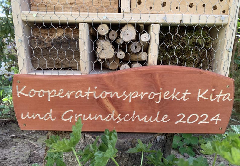 May include: A wooden insect hotel with various natural nesting materials behind wire mesh, featuring a brown wooden sign attached to the front with the engraved white text: Kooperationsprojekt Kita und Grundschule 2024.