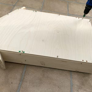 May include: A light-colored wooden structure with a pentagonal top panel being assembled with a power drill. The flat wooden piece is secured to the base frame with multiple small metal screws placed along the edges of the light-toned wood.