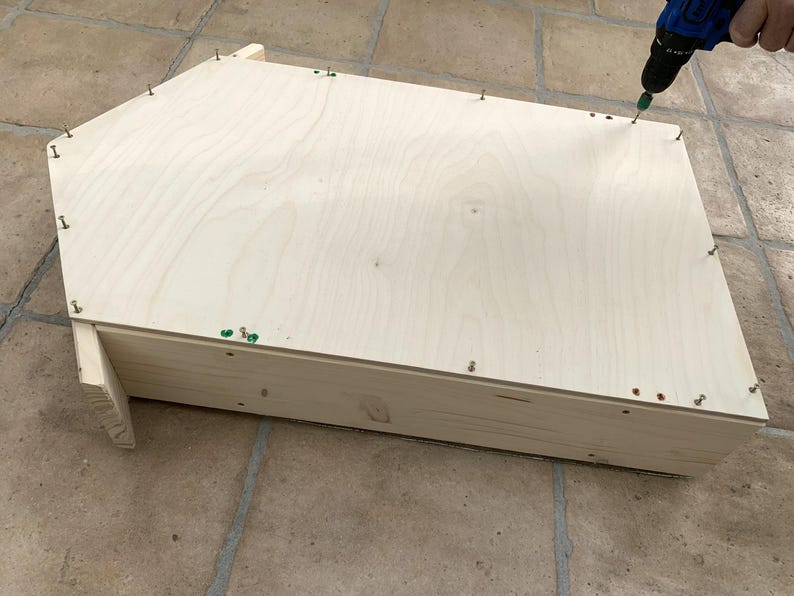 May include: A light-colored wooden structure with a pentagonal top panel being assembled with a power drill. The flat wooden piece is secured to the base frame with multiple small metal screws placed along the edges of the light-toned wood.
