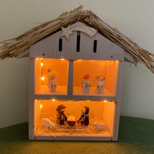 Può includere: Un presepe in legno con tetto di paglia e illuminazione interna. La scena presenta statuine di angeli, pastori e il bambino Gesù in una mangiatoia, con pecore. La combinazione di colori sono toni di legno naturale con luce calda.