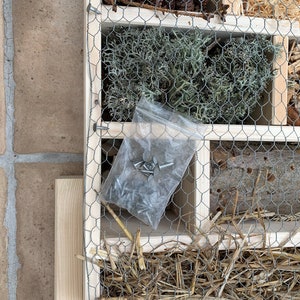 May include: A wooden insect hotel featuring multiple compartments filled with natural materials like pine cones, dried moss, straw, and drilled logs. A hexagonal wire mesh covers the front, and a small plastic bag of metal screws is included.