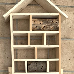 May include: A light-colored wooden insect hotel shaped like a house with a peaked roof. The structure features multiple rectangular compartments of varying sizes, with two cylindrical logs containing drilled holes placed in two of the sections.