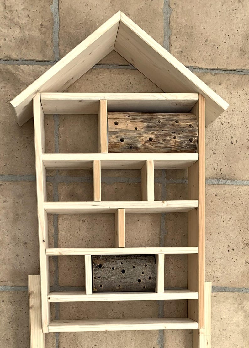 May include: A light-colored wooden insect hotel shaped like a house with a peaked roof. The structure features multiple rectangular compartments of varying sizes, with two cylindrical logs containing drilled holes placed in two of the sections.