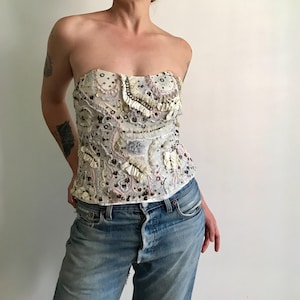 May include: A strapless corset-style top in off-white, featuring intricate embroidery, small ruffles, and scattered sequins in shades of silver, pale pink, and cream. The garment has a structured bodice with a textured, layered surface design.
