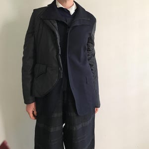 Könnte beinhalten: Eine schwarze und marineblaue Jacke mit einem einzigartigen Design. Die Jacke hat einen gesteppten schwarzen Bereich auf der linken Seite und einen marineblauen Blazer-Stil auf der rechten Seite. Das Model trägt auch eine schwarze Hose.
