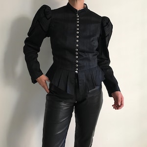 Puede incluir: Una chaqueta negra de manga larga con cuello mao alto, hombros abullonados estructurados con detalles plisados y una hilera de botones de metal plateado en la parte delantera. La prenda incluye un bajo con volante plisado en la cintura.