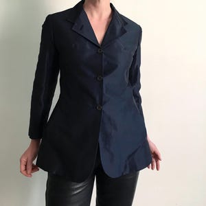 Könnte beinhalten: Ein marineblauer Blazer mit fallendem Revers und Dreiknopfverschluss. Die langärmlige Jacke hat eine taillierte Passform und einen leichten Glanz. Der Blazer wird mit einer schwarzen Lederhose kombiniert.