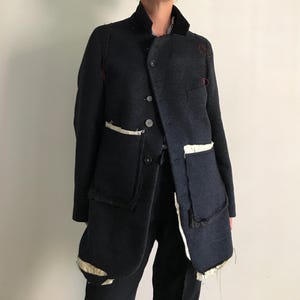 Pode incluir: Um casaco cinza-carvão escuro com gola alta, fecho frontal de botões e dois bolsos grandes com forro branco exposto. A peça apresenta detalhes de costura vermelha visíveis nos ombros e bordas cruas e inacabadas por toda a sua extensão.
