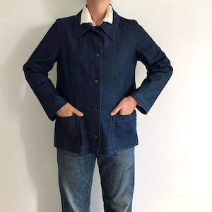 May include: A dark blue denim jacket featuring a pointed collar, long sleeves, and a front button closure with metallic snaps. The jacket includes two large patch pockets on the lower front and is worn over a white collared shirt and blue jeans.