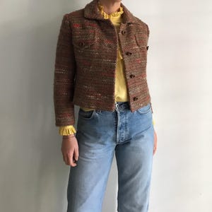 Op de afbeelding: Een bruin tweed jasje met een textuurweefsel en oranje accenten, voorzien van een kraag, knoopsluiting en twee borstzakken. Daaronder een lichtgeel shirt met lange mouwen, een ruchekraag en ruchemanchetten, gecombineerd met een lichtblauwe spijkerbroek.