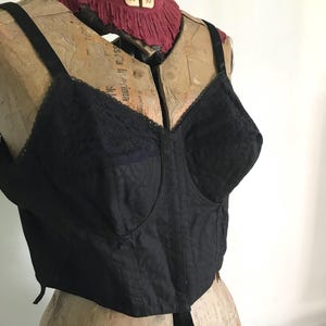May include: A black longline bra featuring a structured bodice with vertical stitching, lace detailing on the upper cups, and adjustable shoulder straps. The garment is displayed on a vintage dress form with a red fabric neck accent.