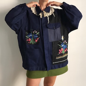Può includere: Una giacca blu navy con colletto bianco a volant, maniche lunghe e chiusura frontale con bottoni. La giacca presenta pannelli in tessuto nero sul davanti con ricami floreali colorati, inclusi fiori blu e rosa, e due tasche sul petto.