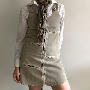 Può includere: Abito in camoscio verde chiaro con rifiniture in crochet bianco e camicia bianca con colletto sotto. L'abito è senza maniche e arriva al ginocchio. Una sciarpa fantasia è indossata intorno al collo.