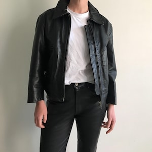 Peut inclure: Une veste en cuir noire avec une fermeture éclair et un col. La veste est portée sur un t-shirt blanc et un pantalon noir. L'ensemble est un style classique et intemporel.