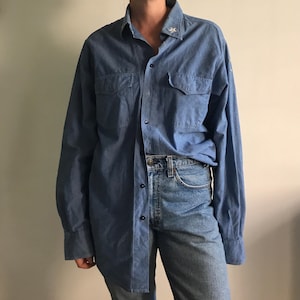 May include: A light blue denim shirt with long sleeves, two chest pockets, and a button-down front. The shirt has a classic collar and a small star-shaped emblem on the collar. The shirt is paired with light blue denim jeans.