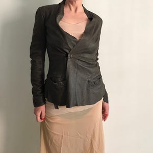 May include: A dark brown leather jacket with a unique asymmetrical design. The jacket features a single button closure, two front pockets, and long sleeves. The jacket is worn over a light peach-colored top and skirt.