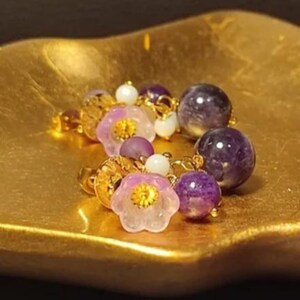 May include: A pair of earrings featuring purple amethyst beads, small white spherical beads, and translucent light purple flower-shaped charms with gold-toned centers. The earrings are constructed with gold-toned metal components and hardware.