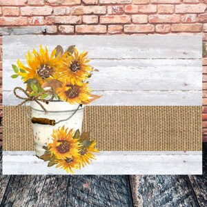 May include: A white metal bucket filled with yellow sunflowers and green leaves. The bucket is sitting on a brown burlap runner and a whitewashed wood background.
