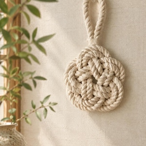 May include: A decorative wall hanging crafted from thick, off-white twisted cotton rope. The rope is intricately woven into a symmetrical, multi-petaled flower shape with a central opening and a sturdy loop at the top for hanging on a wall.