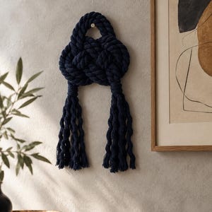 May include: A dark navy blue decorative wall hanging crafted from thick, braided rope. The rope is intricately knotted into a central circular shape with a loop at the top for hanging and two long, dangling tassels extending from the bottom.