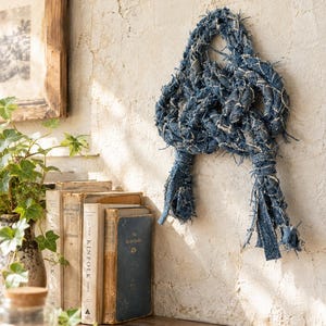 May include: A decorative wall hanging made of braided blue denim fabric strips with frayed edges. The denim is knotted into a complex, intertwined shape with two long, loose ends hanging down, showcasing a textured, worn appearance.