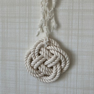May include: A handcrafted, off-white nautical knot wall hanging. The decorative knot is made of thick, twisted rope, suspended from a wooden peg with a frayed, textured rope. The background is a textured, light-colored wall.