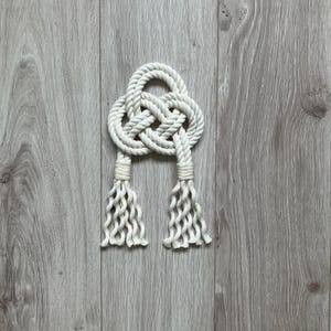 May include: A white rope knot with tassels, tied in a Celtic knot design. The knot is on a wood background.