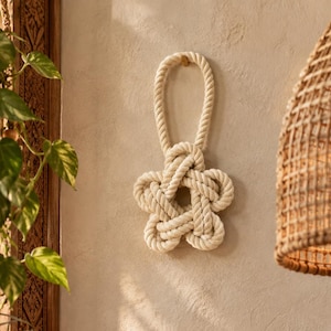 May include: A wall-mounted decorative object crafted from thick, off-white twisted rope. The rope is intricately knotted into a five-pointed star shape with a large loop at the top for hanging, creating a textured, nautical-inspired design.